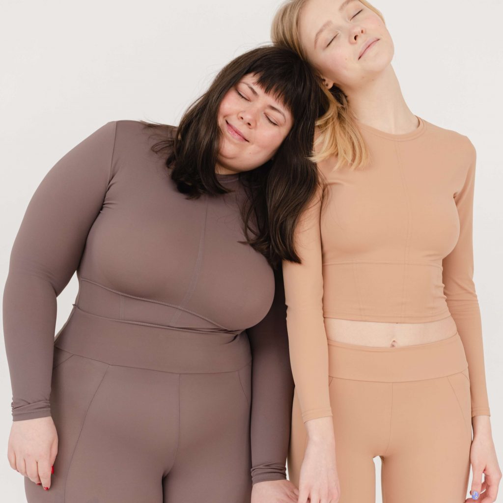 two women standing next to each in skin tight clothing, resting heads on each other
